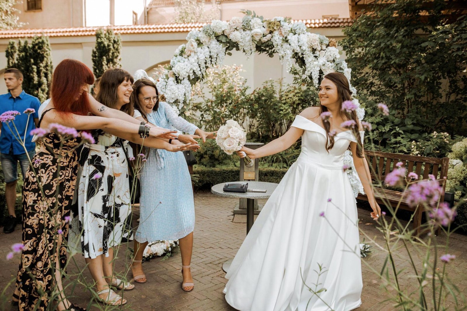 A bride in a white gown holds a bouquet while several women reach out to catch it in a garden setting. The background features an arch decorated with white flowers and trees, creating a picturesque scene. The atmosphere is festive and joyful with blooming flowers in the foreground.