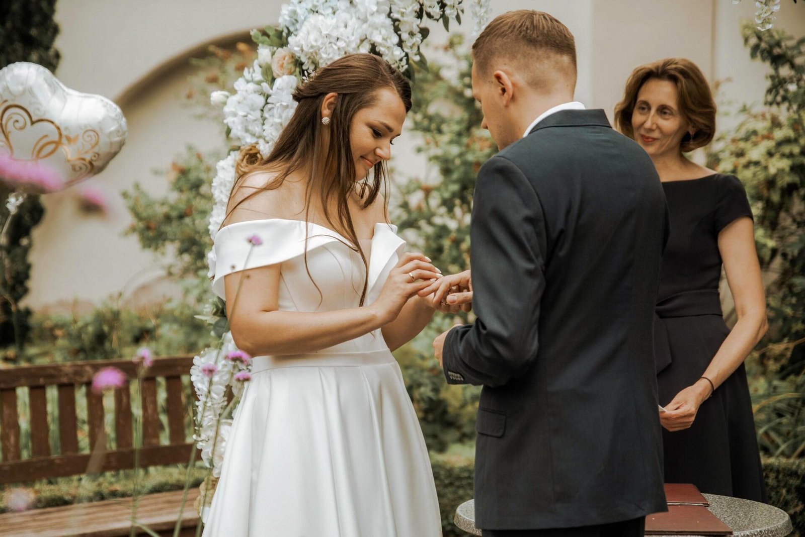 A bride in an off-shoulder white gown exchanges rings with the groom, who is in a dark suit, during an outdoor wedding ceremony. Under an arch decorated with flowers, a woman in a black dress officiates. A heart-shaped balloon is visible in the background—captured perfectly by a luxury wedding photographer in Marbella.