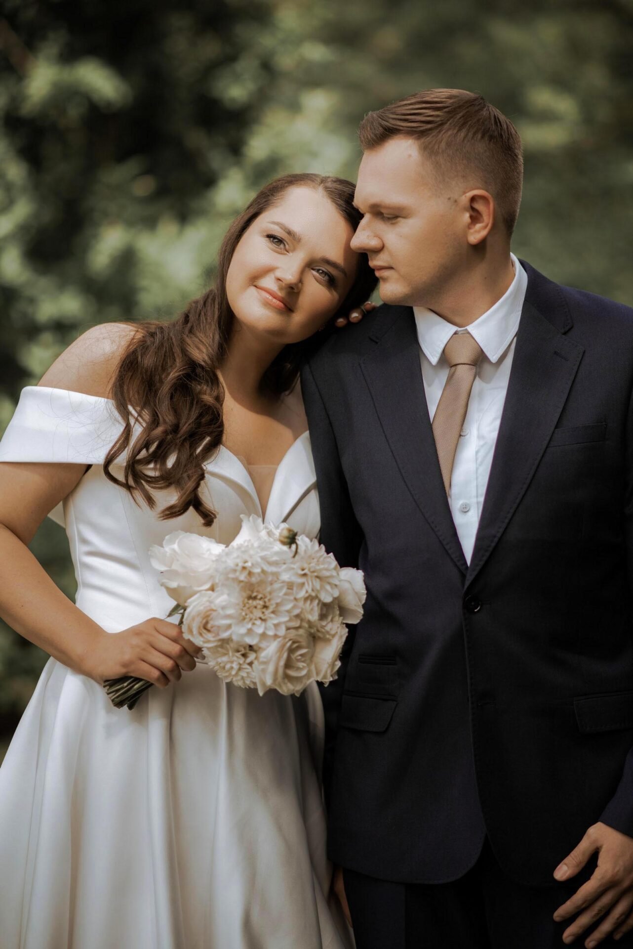 A bride and groom stand closely together in an outdoor setting. The bride, wearing an off-shoulder white gown and holding a bouquet of white flowers, smiles while leaning her head on the groom's shoulder. The groom is dressed in a dark suit with a white shirt and tan tie. Captured beautifully by a luxury wedding photographer in Marbella.