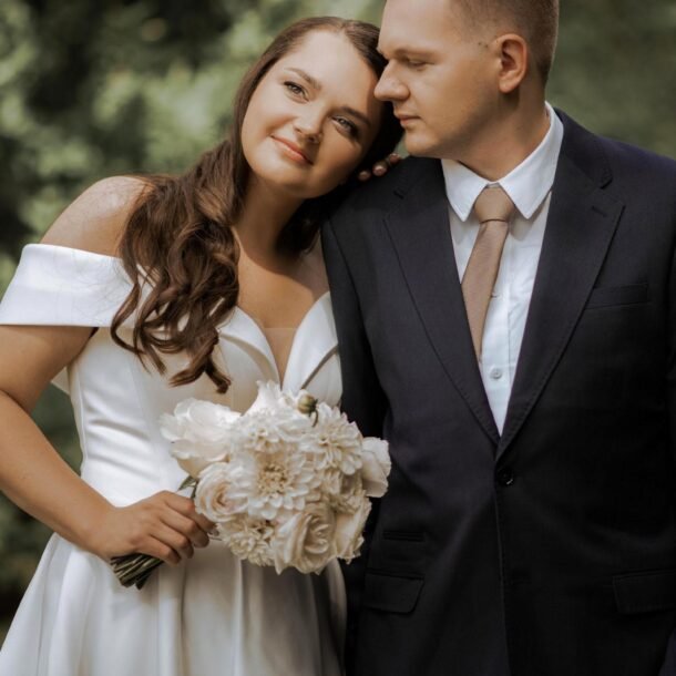 A bride and groom stand closely together in an outdoor setting. The bride, wearing an off-shoulder white gown and holding a bouquet of white flowers, smiles while leaning her head on the groom's shoulder. The groom is dressed in a dark suit with a white shirt and tan tie. Captured beautifully by a luxury wedding photographer in Marbella.