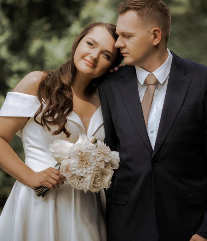 A bride and groom stand closely together in an outdoor setting. The bride, wearing an off-shoulder white gown and holding a bouquet of white flowers, smiles while leaning her head on the groom's shoulder. The groom is dressed in a dark suit with a white shirt and tan tie. Captured beautifully by a luxury wedding photographer in Marbella.