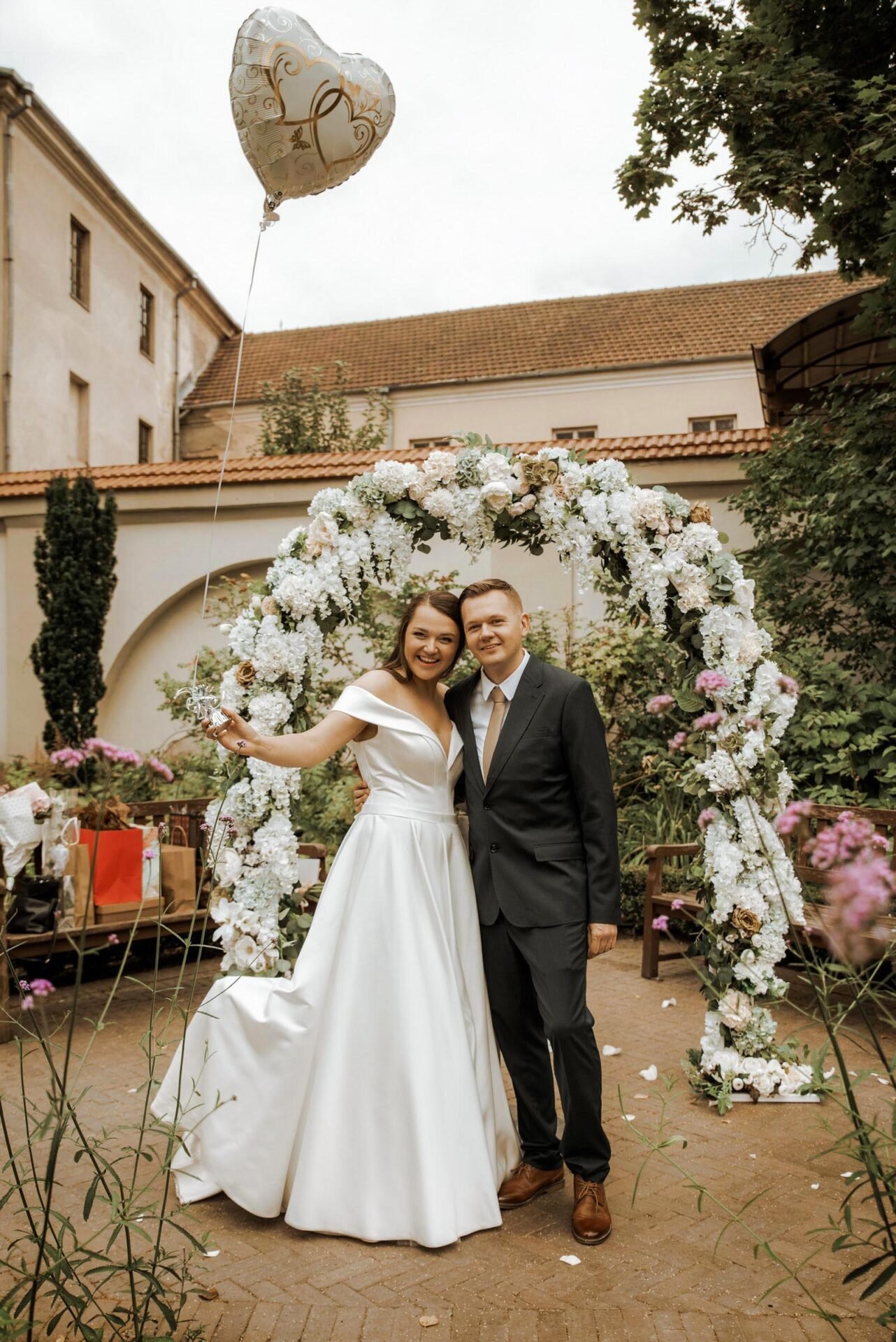 A bride in a white wedding dress and a groom in a black suit stand smiling under a floral arch decorated with white flowers. The bride holds a heart-shaped balloon. They are outdoors, surrounded by flowers and greenery, with buildings in the background—a perfect scene for wedding photography Marbella.