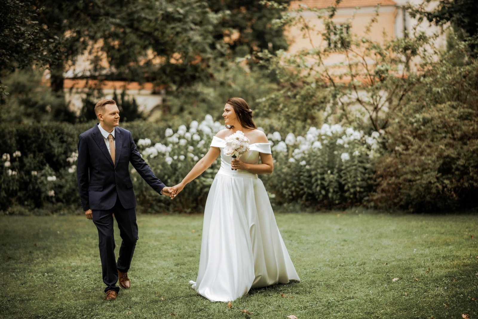 A bride and groom walk hand-in-hand in a garden. The bride is wearing a long, white off-the-shoulder dress and holding a bouquet of white flowers. The groom is dressed in a dark suit with a tie. They are surrounded by greenery and white flowers, captured beautifully by their luxury wedding photographer in Marbella.