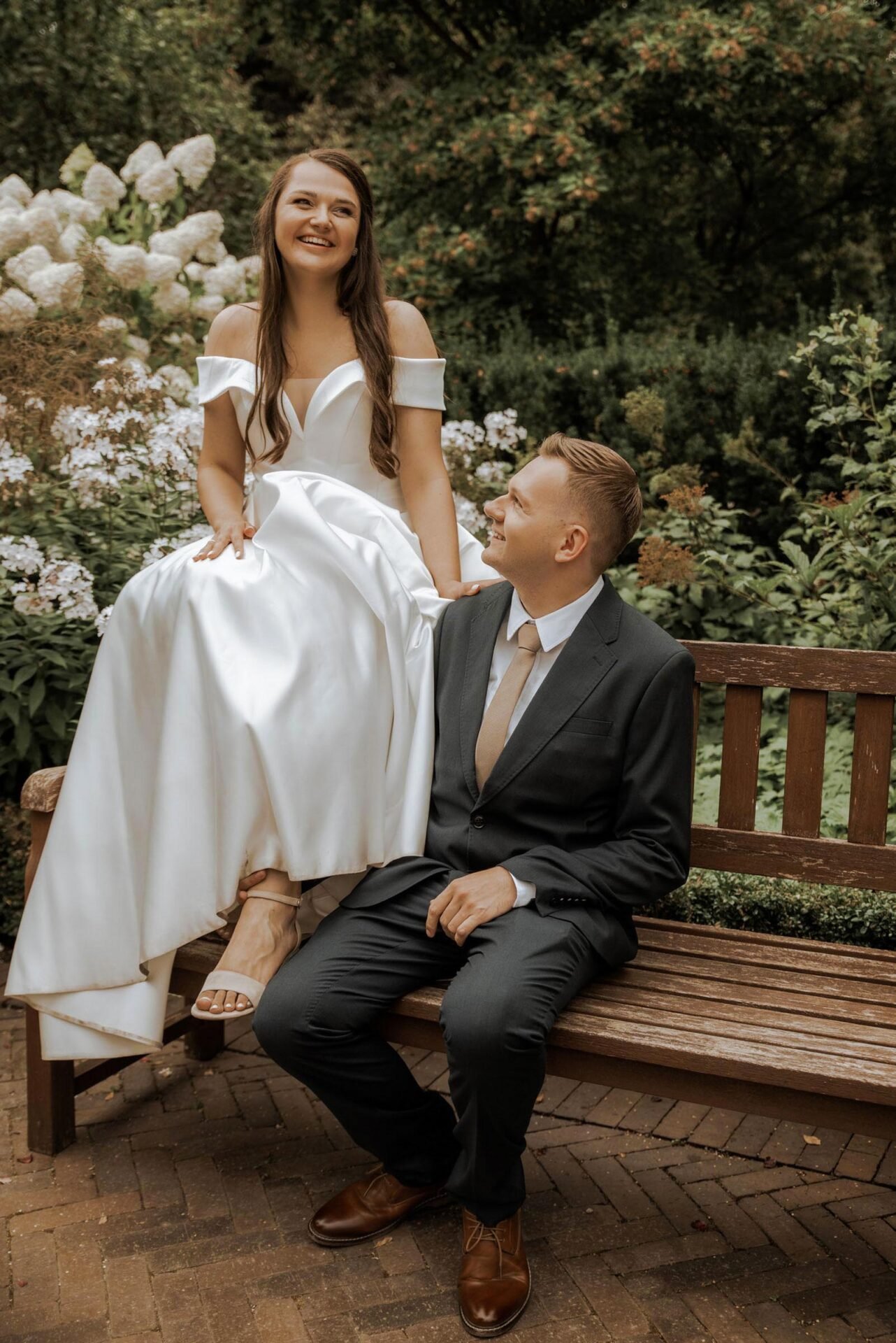 A bride in a white gown sits on the backrest of a wooden bench, smiling and looking away. The groom, in a dark suit, sits on the bench seat and gazes up at the bride. They are surrounded by lush greenery and white flowers in an outdoor garden setting.