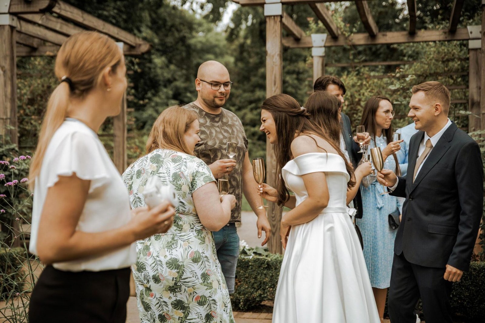 A group of people socializing outdoors. A woman in a white off-the-shoulder dress laughs while holding a champagne flute. Two men and several women with drinks in hand engage in conversation. The scene, perfect for a wedding photographer Costa del Sol, is set under a wooden structure with greenery in the background.