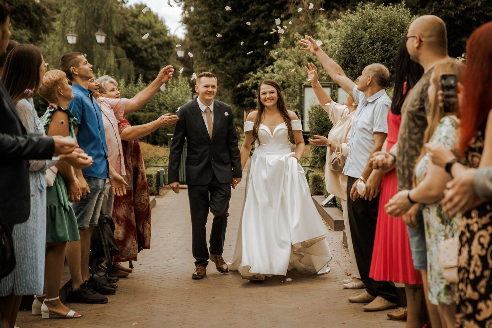 A bride and groom walk hand-in-hand down a path, smiling. Surrounded by friends and family tossing flower petals in celebration, the bride wears a flowing white gown while the groom is in a dark suit and tie. Trees and greenery form the perfect backdrop for this Luxury Wedding Photographer Marbella scene.