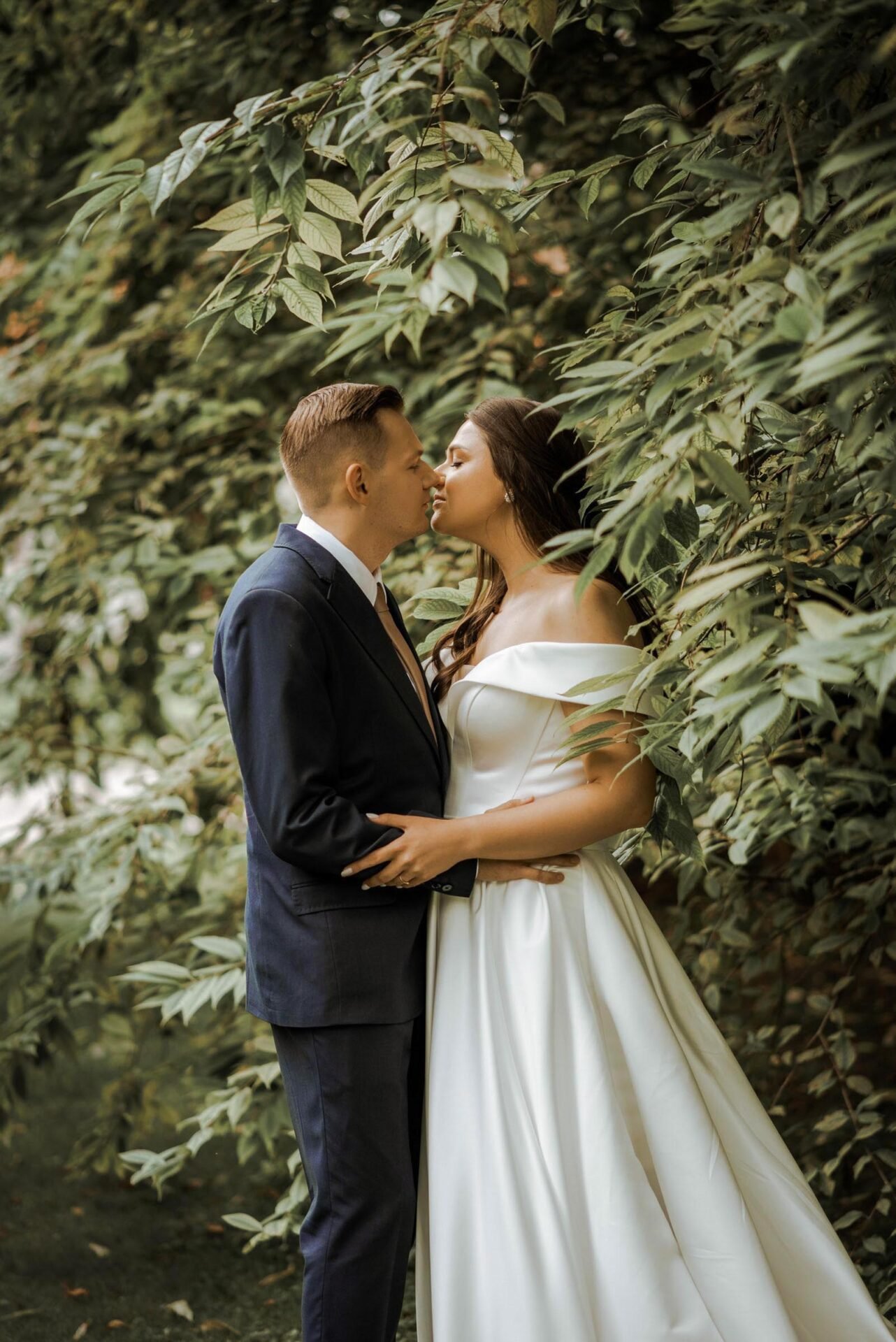 A couple dressed in wedding attire stands close, gazing into each other's eyes. The woman wears an off-shoulder white wedding gown, and the man is in a dark suit. They are partially surrounded by lush, green foliage, creating a romantic and intimate setting captured perfectly by their Destination Wedding Photographer Marbella.