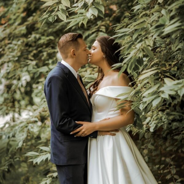 A couple dressed in wedding attire stands close, gazing into each other's eyes. The woman wears an off-shoulder white wedding gown, and the man is in a dark suit. They are partially surrounded by lush, green foliage, creating a romantic and intimate setting captured perfectly by their Destination Wedding Photographer Marbella.