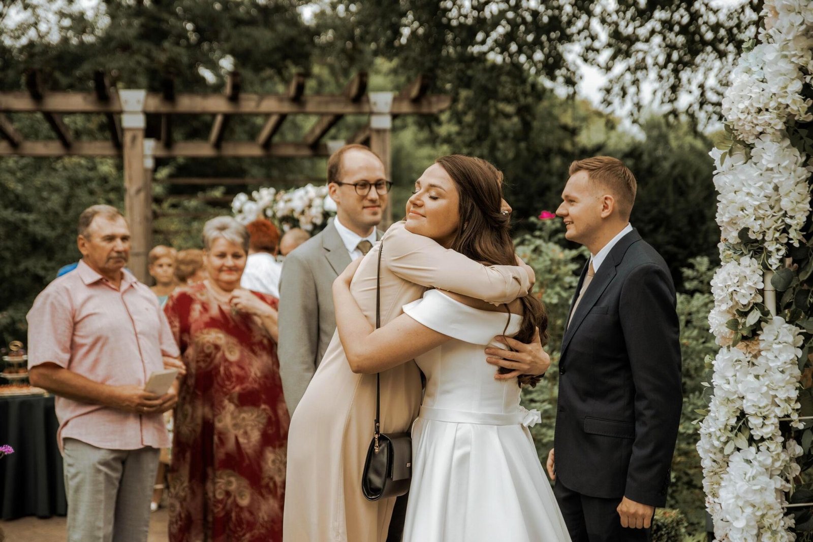 A bride in a white dress is embracing a guest at an outdoor wedding ceremony. The groom and more guests are standing nearby. There is greenery and floral decorations around, with a wooden pergola in the background, capturing the essence of Wedding Photography Marbella.