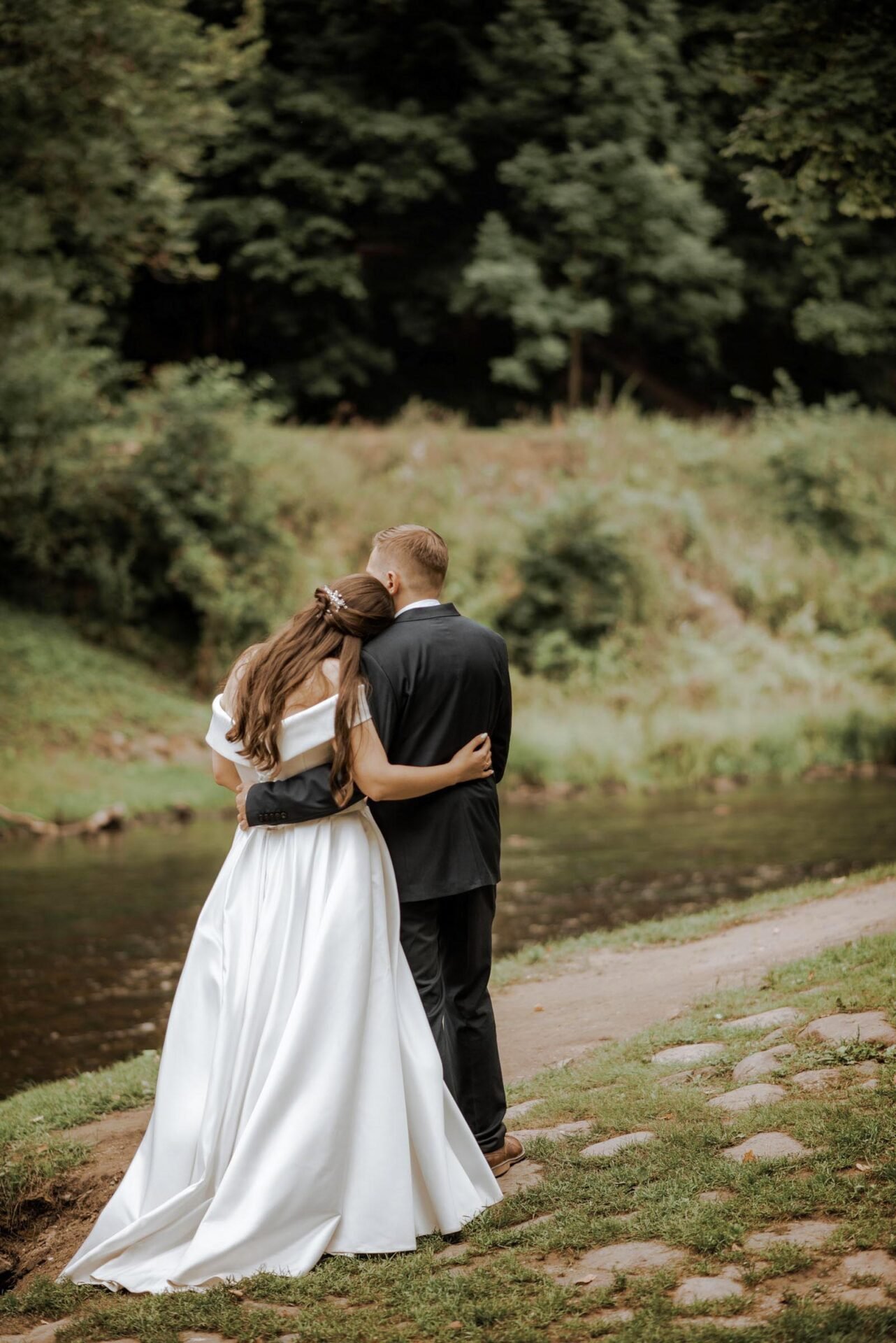 A couple in wedding attire stands by a river with their backs to the camera. The bride wears a white gown with a train, and the groom is in a black suit. They embrace each other, looking towards the lush greenery across the water, captured beautifully by their Wedding Photographer Costa del Sol.