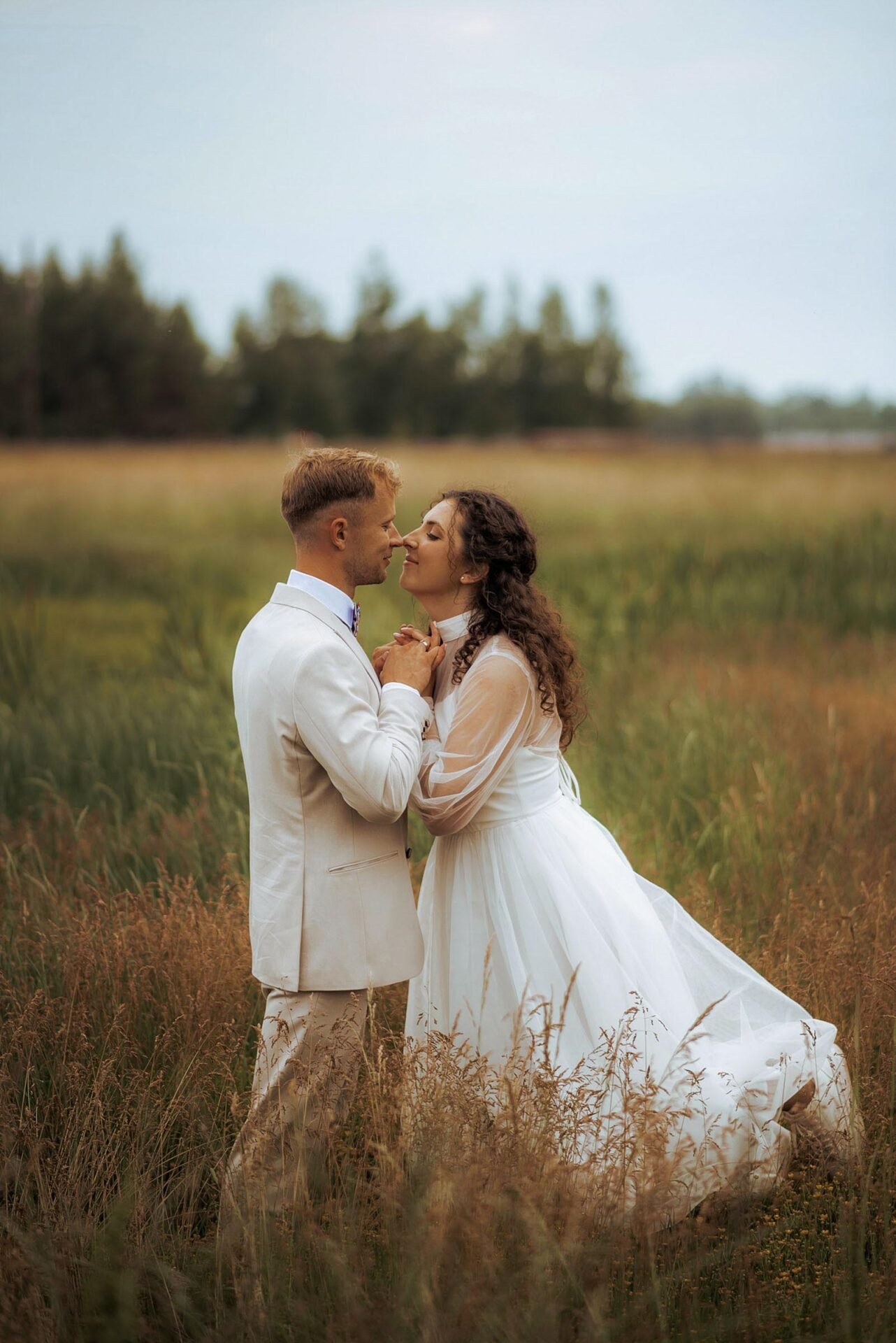 A couple stands in a grassy field, gazing into each other's eyes. The woman wears a long white dress with sheer sleeves, while the man is dressed in a light-colored suit. Captured by a Wedding Photography Marbella expert, the blurred outdoor setting of trees and an overcast sky adds to the romantic charm.