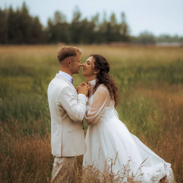 A couple stands in a grassy field, gazing into each other's eyes. The woman wears a long white dress with sheer sleeves, while the man is dressed in a light-colored suit. Captured by a Wedding Photography Marbella expert, the blurred outdoor setting of trees and an overcast sky adds to the romantic charm.