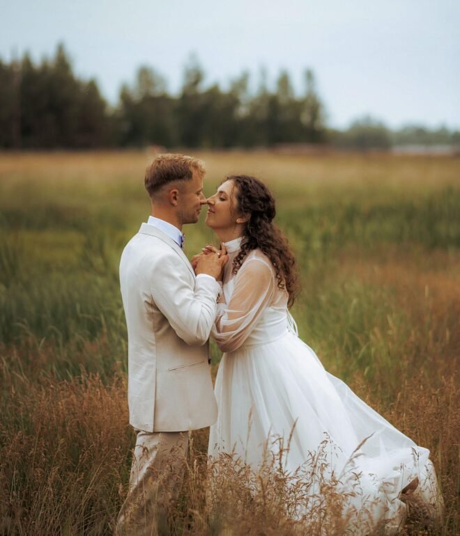 A couple stands in a grassy field, gazing into each other's eyes. The woman wears a long white dress with sheer sleeves, while the man is dressed in a light-colored suit. Captured by a Wedding Photography Marbella expert, the blurred outdoor setting of trees and an overcast sky adds to the romantic charm.