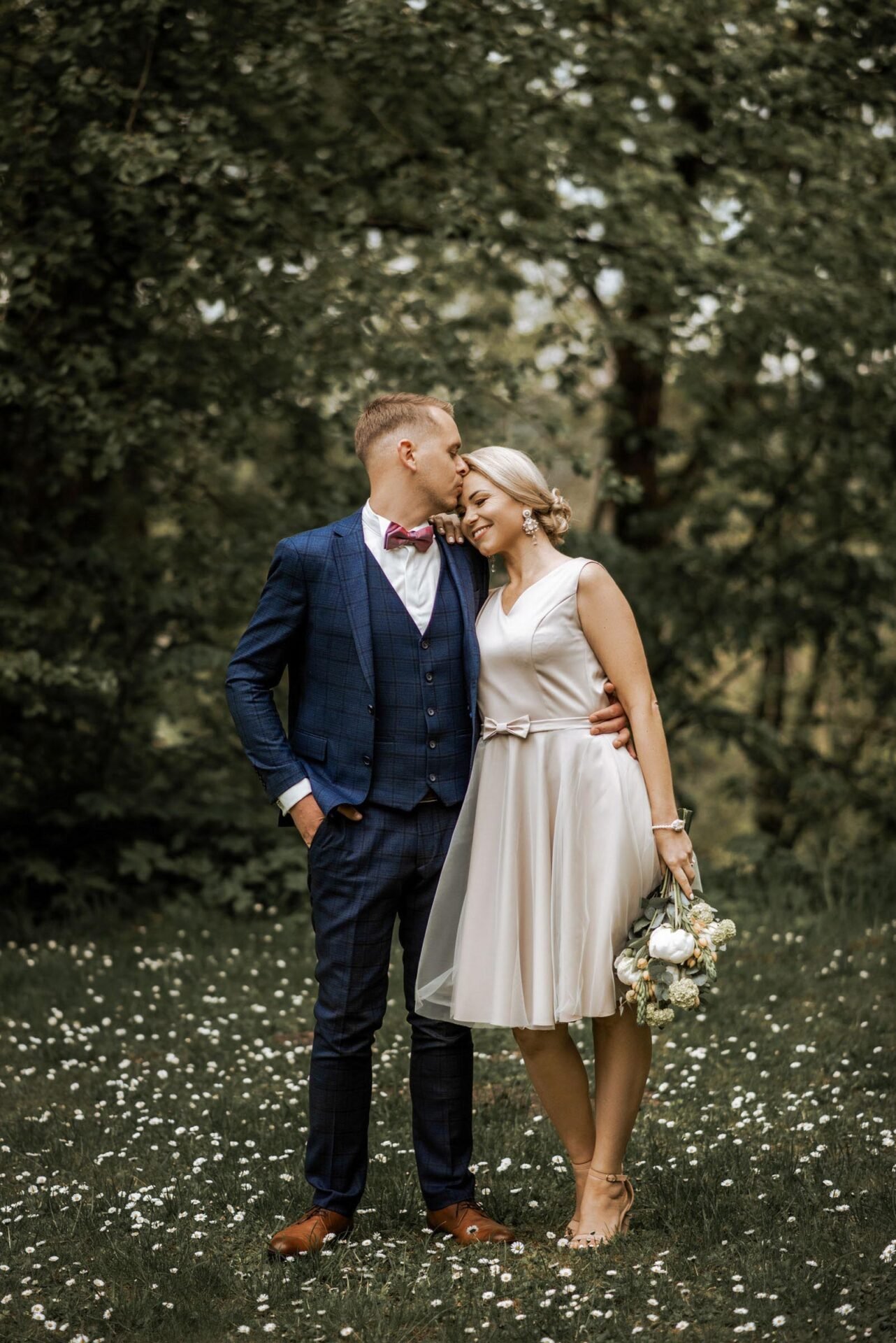 A couple stands intimately on a grassy field with wildflowers. The man, in a navy blue suit with a pink bow tie, kisses the smiling woman, who is wearing a short, light-colored dress and holding a bouquet of flowers. Trees fill the background, creating a serene scene perfect for Wedding Photography Marbella.