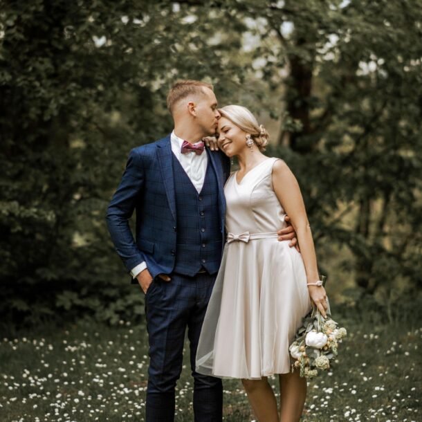 A couple stands intimately on a grassy field with wildflowers. The man, in a navy blue suit with a pink bow tie, kisses the smiling woman, who is wearing a short, light-colored dress and holding a bouquet of flowers. Trees fill the background, creating a serene scene perfect for Wedding Photography Marbella.