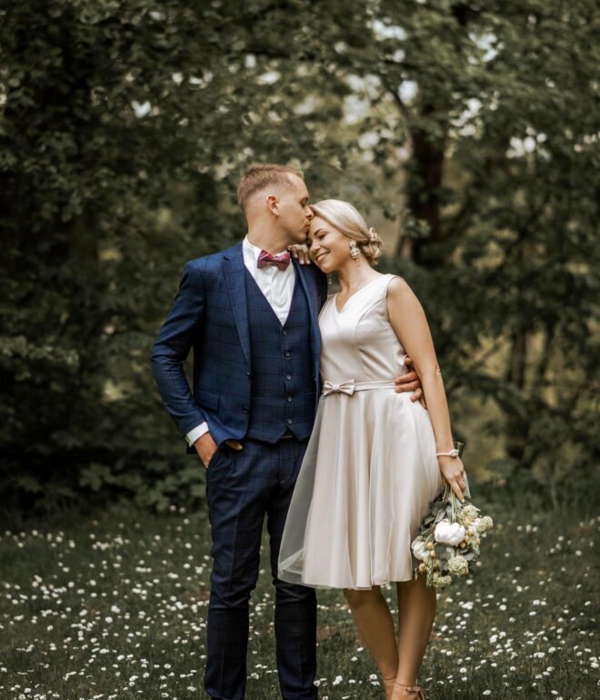 A couple stands intimately on a grassy field with wildflowers. The man, in a navy blue suit with a pink bow tie, kisses the smiling woman, who is wearing a short, light-colored dress and holding a bouquet of flowers. Trees fill the background, creating a serene scene perfect for Wedding Photography Marbella.