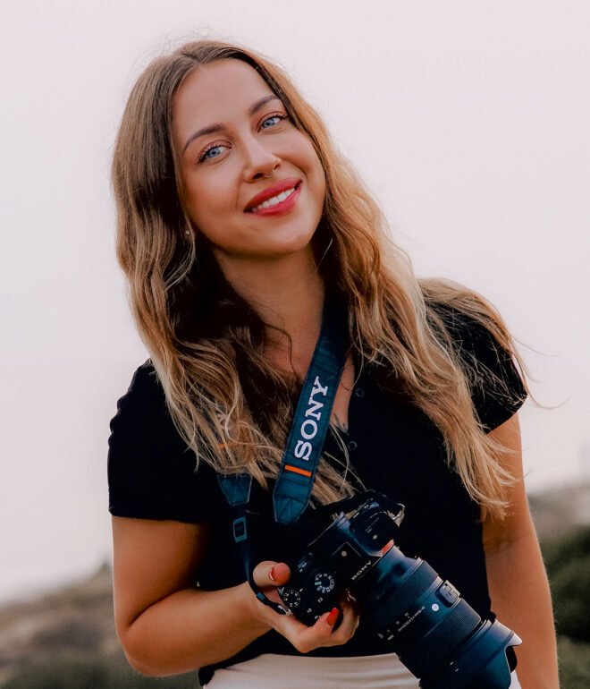 A woman with long wavy hair is smiling and holding a camera. She is wearing a black top and has a camera strap labeled "Sony" around her neck. The background is a scenic outdoor setting with a soft focus on natural scenery.