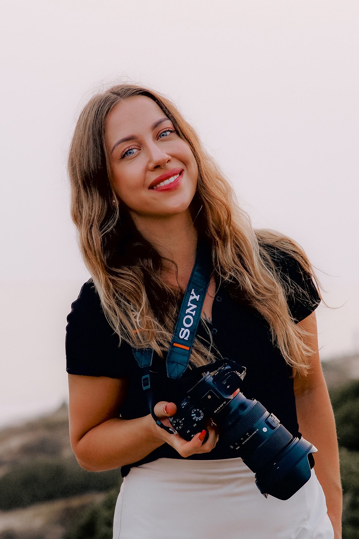 A woman with long wavy hair is smiling and holding a camera. She is wearing a black top and has a camera strap labeled "Sony" around her neck. The background is a scenic outdoor setting with a soft focus on natural scenery.