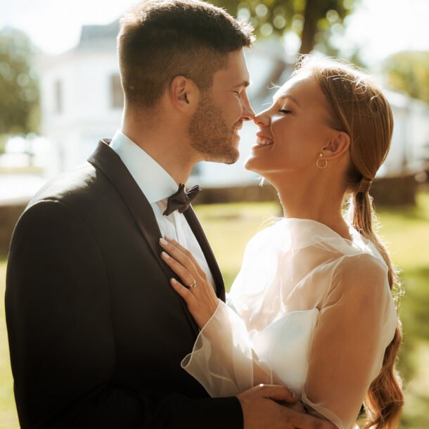 A bride and groom share a joyful moment outdoors. The groom is in a black suit and bow tie, and the bride is in a white gown with sheer sleeves. They are close, smiling, with sunlit greenery in the background.