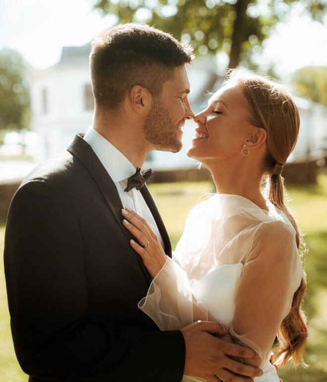 A bride and groom share a joyful moment outdoors. The groom is in a black suit and bow tie, and the bride is in a white gown with sheer sleeves. They are close, smiling, with sunlit greenery in the background.