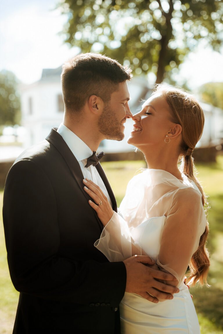 A bride and groom share a joyful moment outdoors. The groom is in a black suit and bow tie, and the bride is in a white gown with sheer sleeves. They are close, smiling, with sunlit greenery in the background.