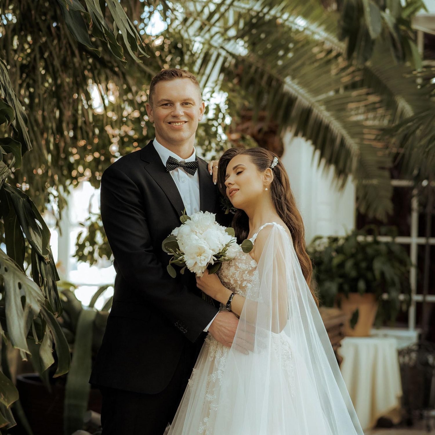 A groom in a black suit and bow tie stands amidst lush greenery, smiling warmly while holding a bouquet. The bride in a white wedding dress with a long veil embraces him affectionately, her eyes closed and smiling, creating a serene and loving scene captured perfectly by the Marbella photographer.