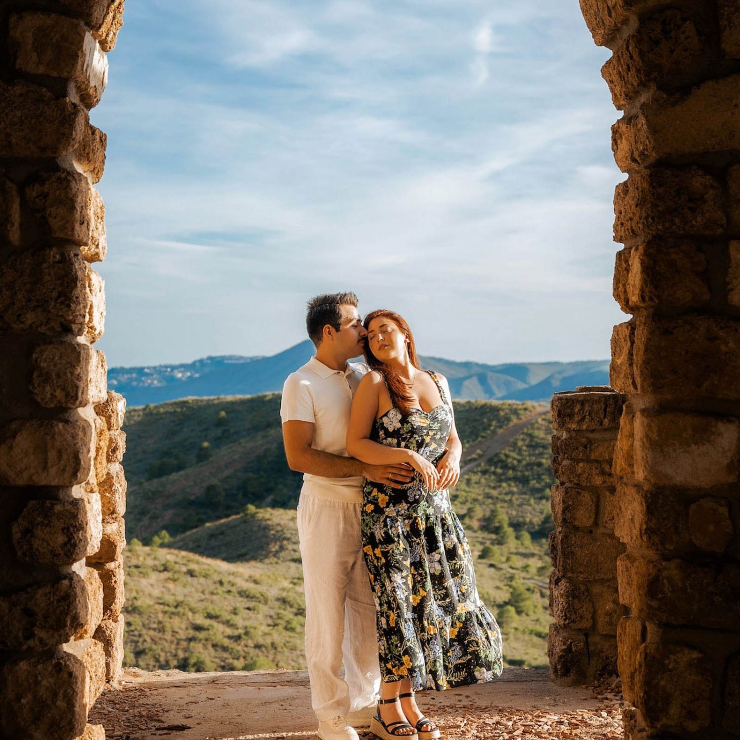 A couple stands in a stone archway overlooking a vast landscape of hills, captured perfectly during their couple photoshoot. The man, dressed in a white shirt and pants, hugs the woman from behind. The woman, in a floral dress, cradles her baby bump as they both gaze into the distance with serene expressions.