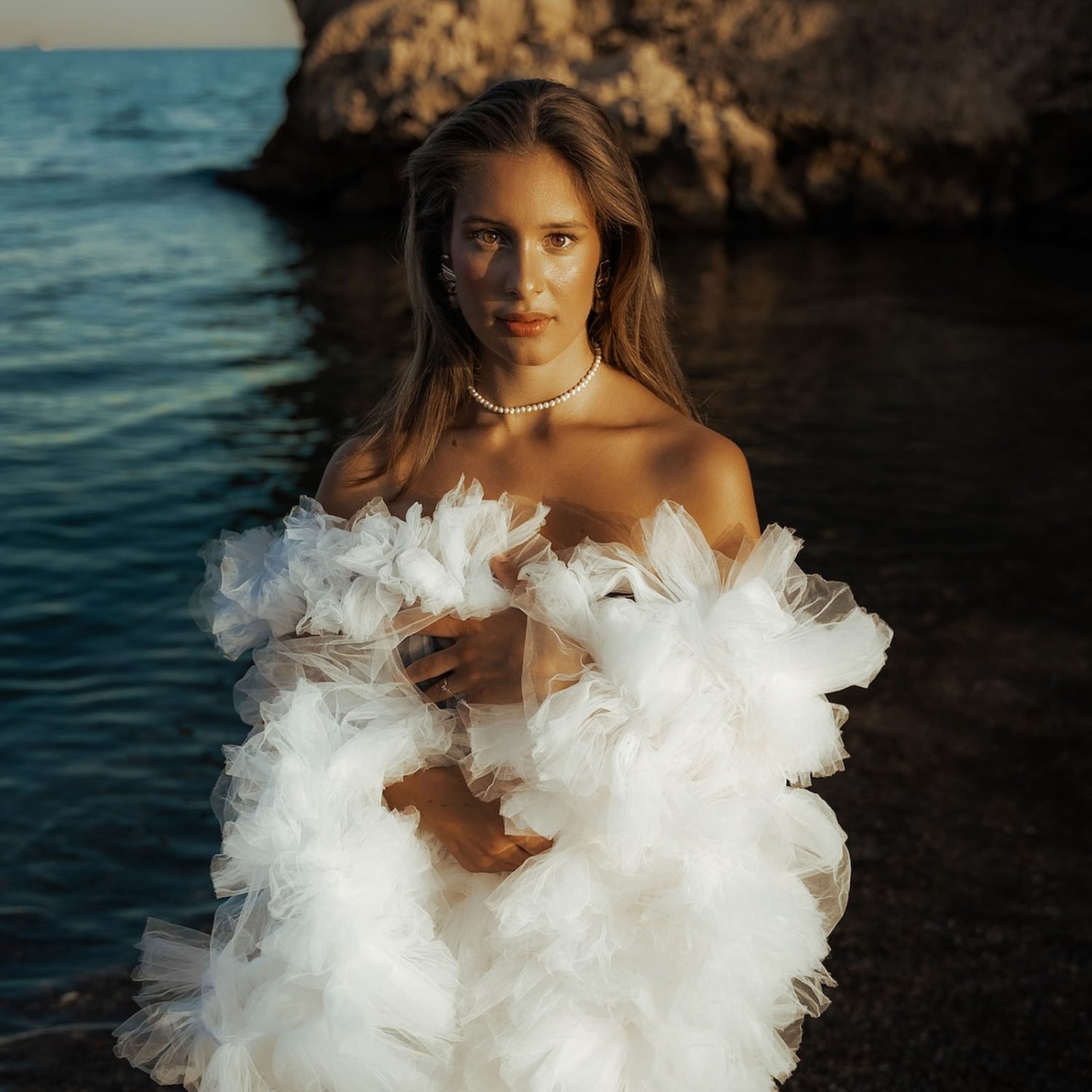 A woman stands by the ocean in an elaborate white tulle gown, her hair glowing in the golden sunlight. This ethereal photoshoot in Costa Del Sol captures the beauty of the seaside.